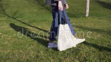 一个穿牛仔裤的女人手里拿着一个米色的棉包，在草坪上的公园里散步。 天然<strong>环保袋</strong>